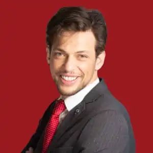 Un hombre sonriente de pelo castaño, con traje oscuro, camisa blanca y corbata roja, de pie sobre un fondo rojo sólido, mirando a la cámara con los brazos cruzados.